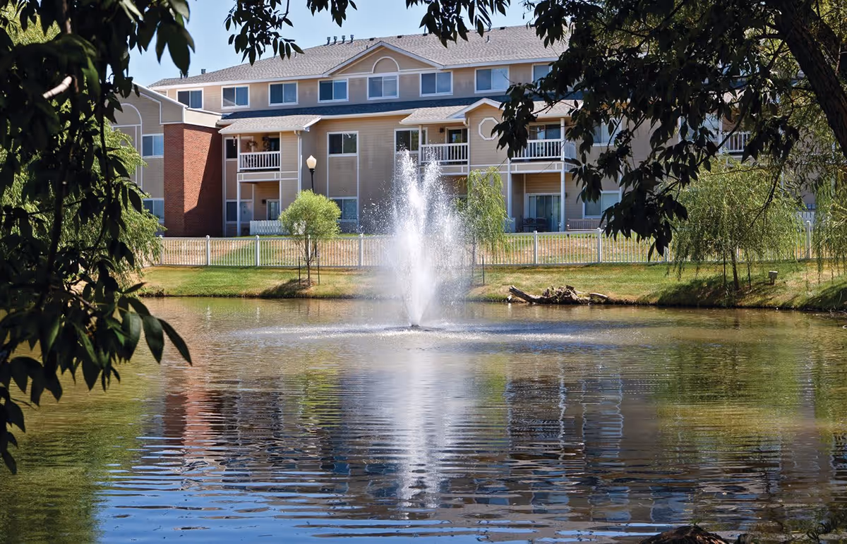 A serene outdoor scene at The Fountains at Greenbriar featuring a pond with a central water fountain, surrounded by green grass, small trees, and a beige multi-story residential building in the background. The image is framed by leafy tree branches.