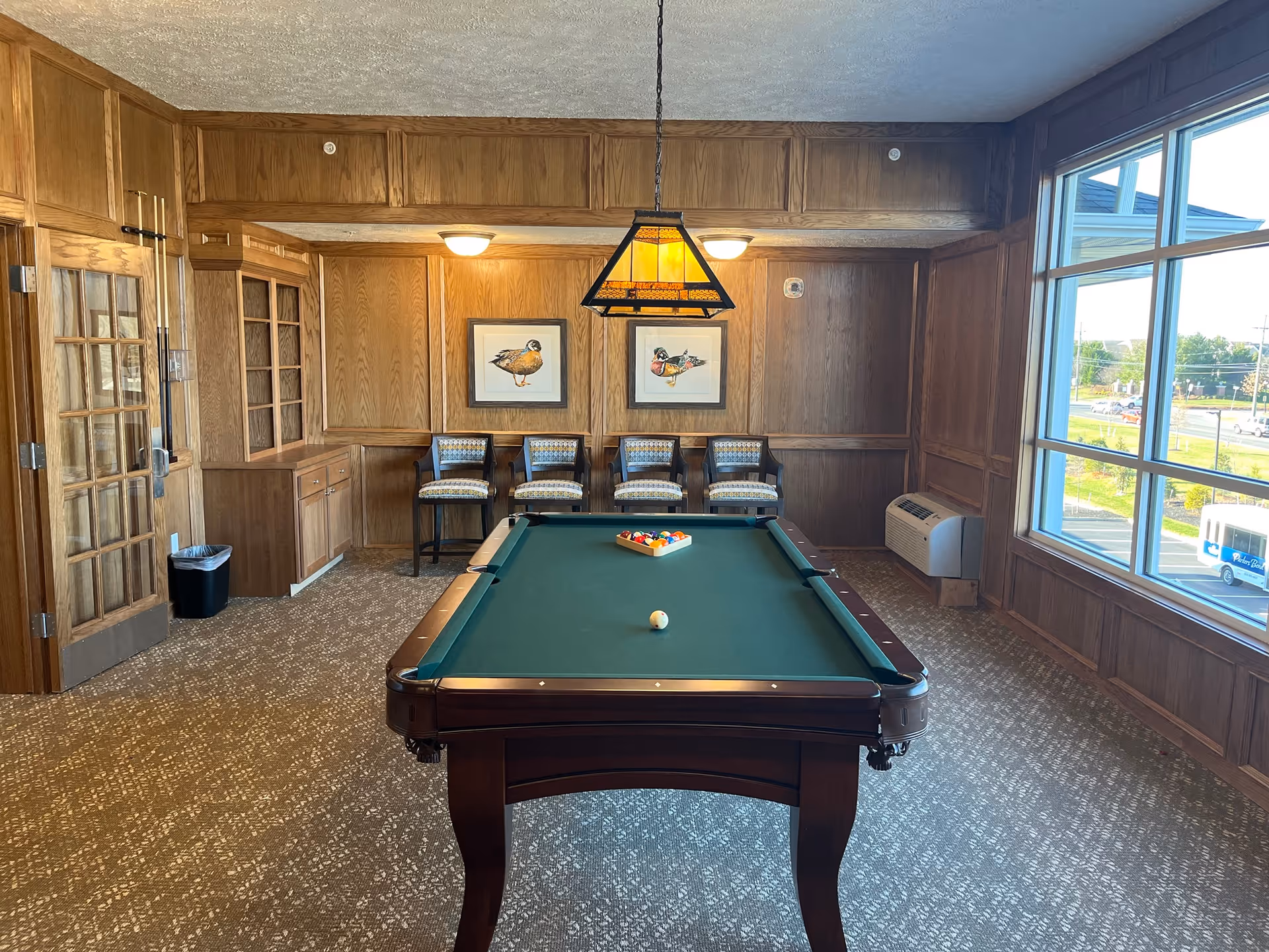A room with wood-paneled walls and carpeted floor featuring a pool table in the center with billiard balls arranged on it. Four cushioned chairs are lined up against the back wall beneath two framed bird paintings. A hanging light fixture is above the pool table, and large windows on the right side let in natural light, showing a view of the outside parking area and street.