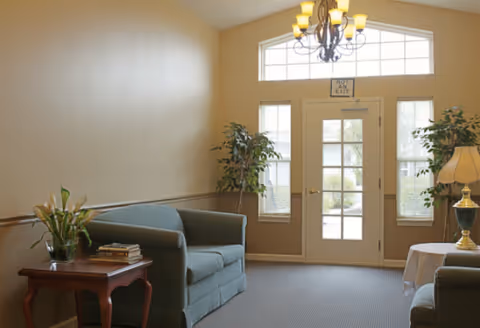 A cozy living room area with two green upholstered sofas facing each other, a wooden side table with a vase of flowers and books, a round table with a lamp, and a large windowed door letting in natural light. There are two potted plants near the door and a chandelier hanging from the ceiling.