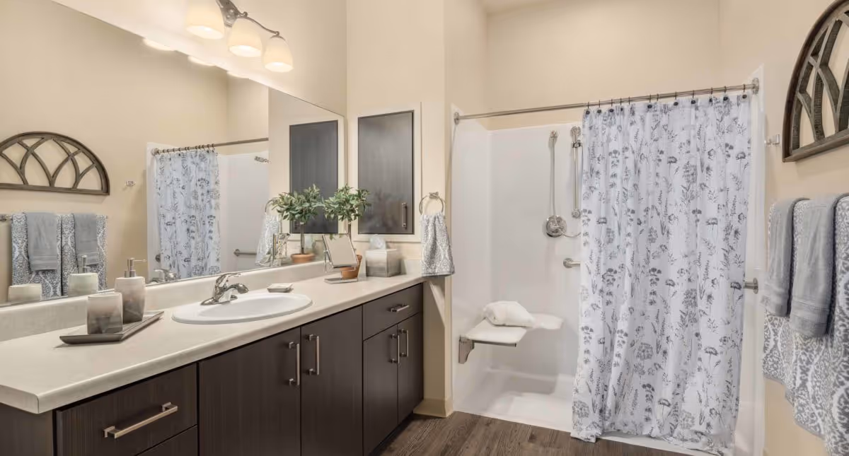 Accessible bathroom with a long vanity and sink, large mirror, and a walk-in shower with a patterned curtain and fold-down seat.