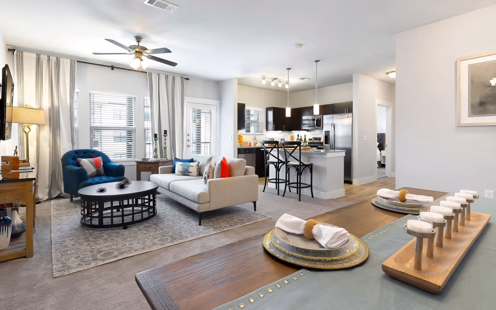 A bright and modern open-concept living space featuring a living room with a beige sofa, blue armchair, round black coffee table, and a TV mounted on the wall. The living room flows into a kitchen area with dark cabinets, stainless steel appliances, a kitchen island with two black bar stools, and pendant lighting. In the foreground, a dining table is set with plates, napkins, and a decorative candle holder. Large windows with striped curtains allow natural light to fill the room.