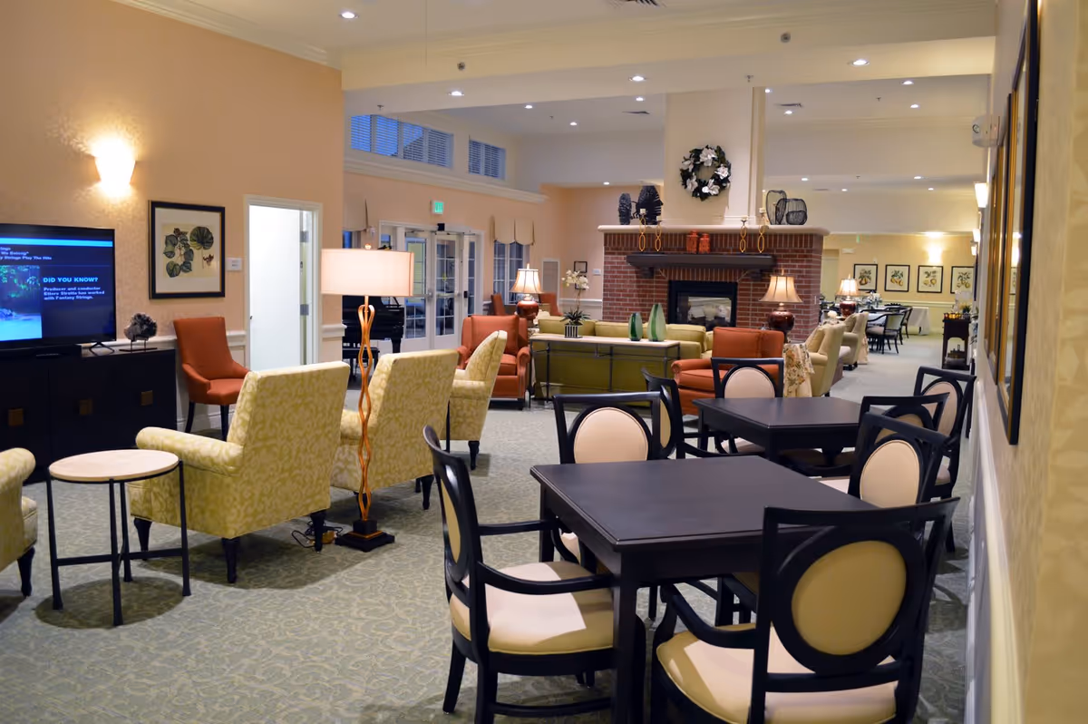 A spacious senior living common area with multiple seating arrangements including cushioned armchairs, sofas, and dining tables with chairs. The room features a brick fireplace decorated with a wreath and various ornaments. There are lamps providing warm lighting, framed artwork on the walls, and a television on a cabinet. The space has a carpeted floor and light-colored walls, creating a cozy and inviting atmosphere.