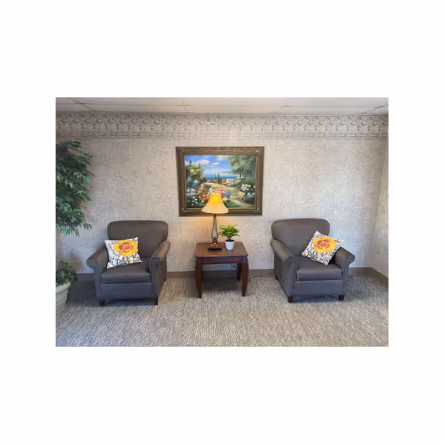 Two upholstered armchairs flank a small wooden side table with a lamp beneath a framed landscape painting in a waiting-area style interior.