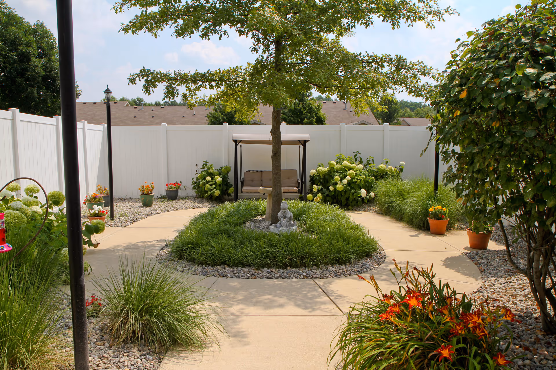 Sunlit fenced courtyard garden with a central tree surrounded by grass, a paved circular walkway, potted flowers and a covered swing bench.