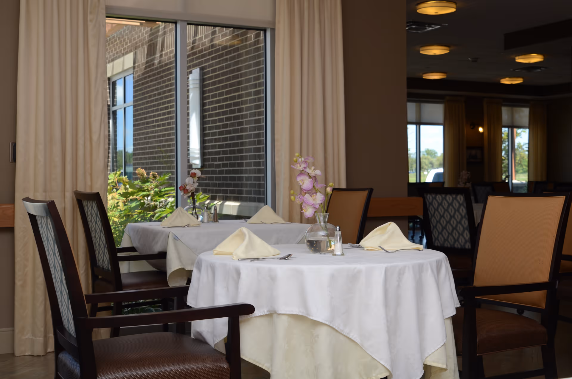 A dining area with tables covered in white tablecloths, each set with folded napkins, silverware, and a small vase with flowers. The room has large windows with beige curtains, and outside greenery is visible through the windows.