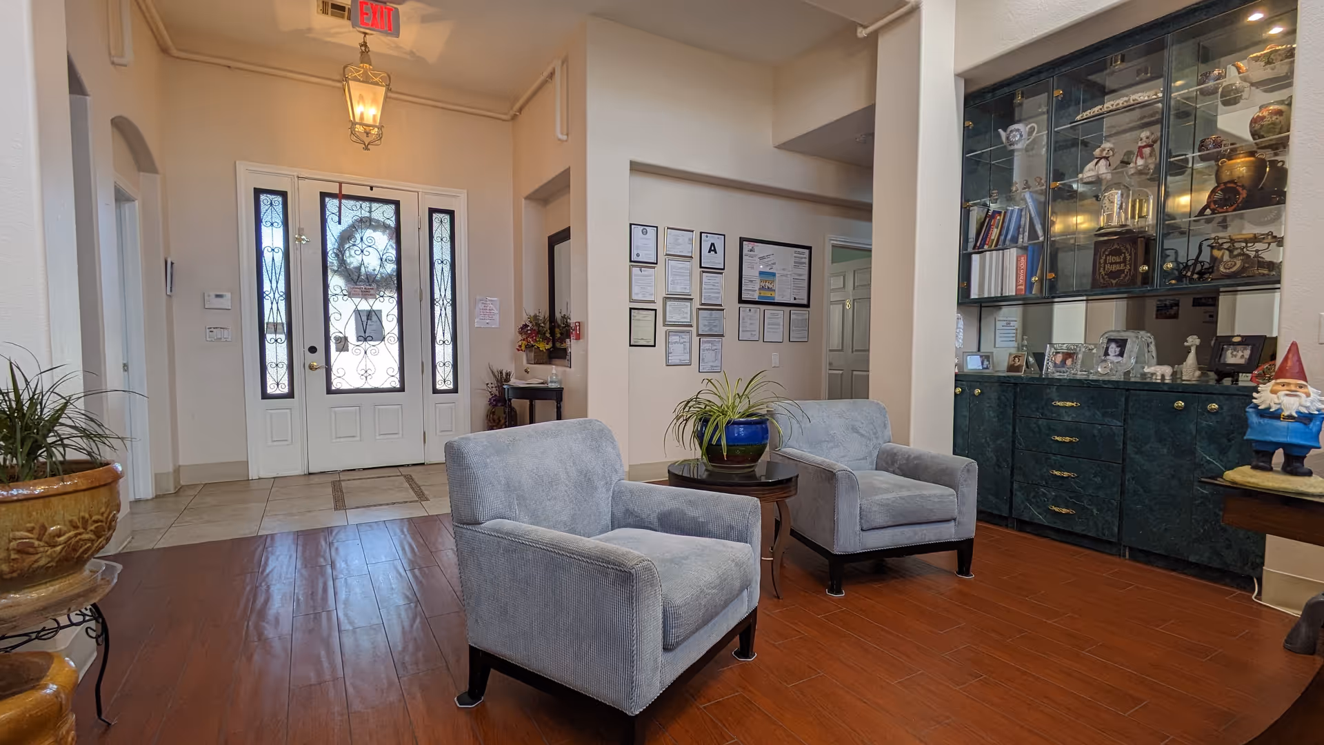 Entrance lobby of a senior home with two upholstered chairs around a small table, front double doors, and a glass display cabinet.