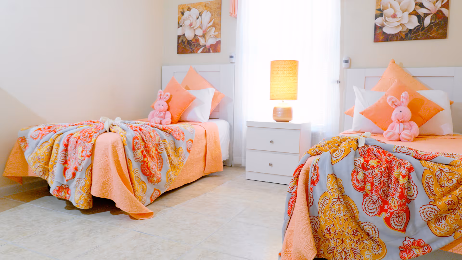 A bright bedroom with two twin beds, each adorned with orange and patterned bedspreads, orange pillows, white pillows, and a pink stuffed bunny. Between the beds is a white nightstand with two drawers and a lit table lamp. The walls are light-colored with floral paintings above each bed, and a window with sheer white curtains is behind the nightstand.