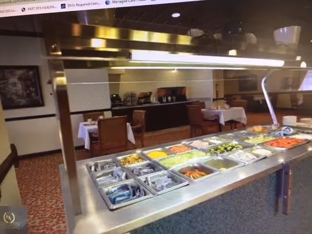 Buffet-style food serving area with various trays of food including vegetables, fruits, and packaged items, set in a dining room with tables and chairs in the background.