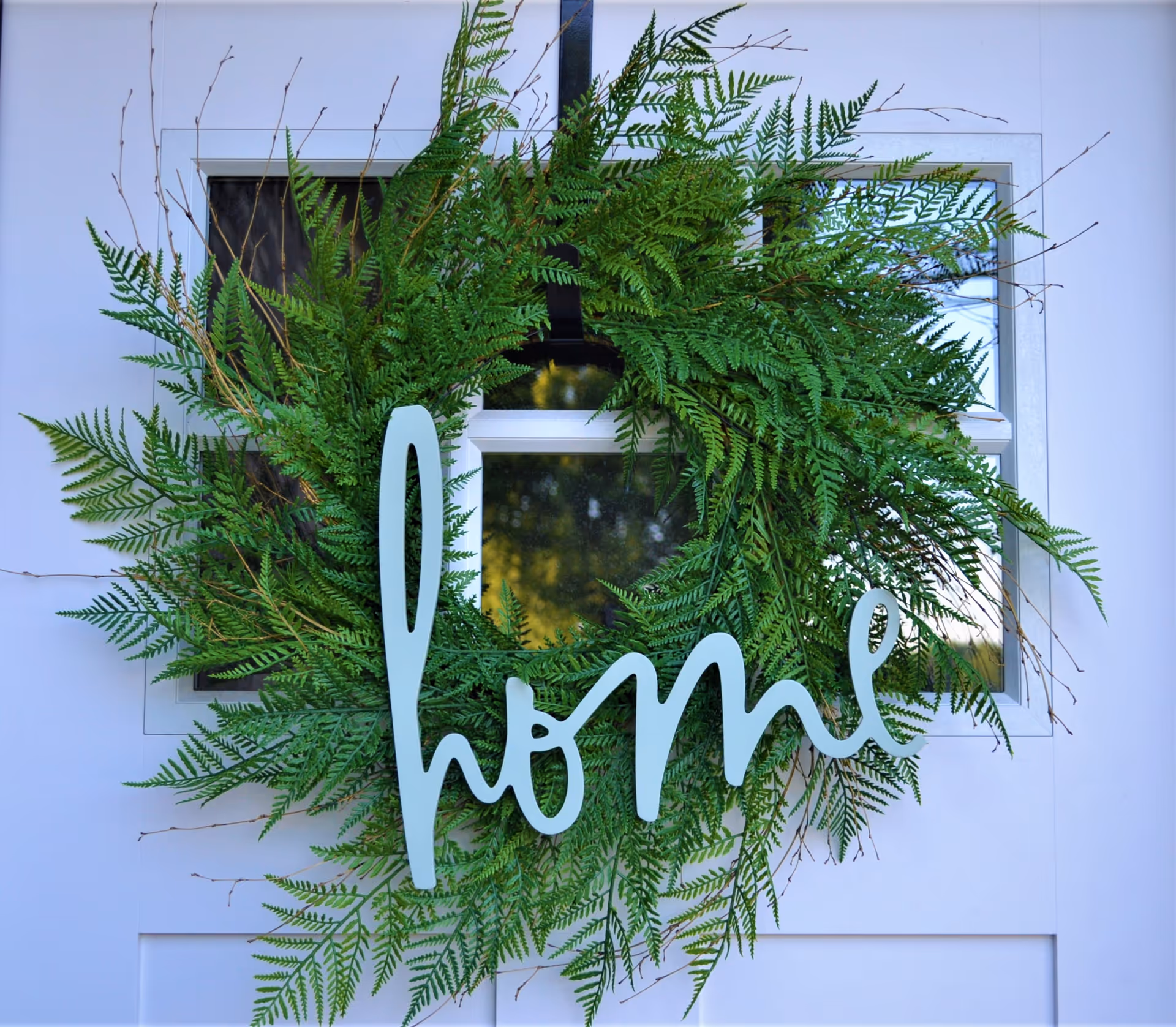 A green fern wreath hanging on a white door with a window. The wreath has a decorative word 'home' attached to it in light blue color.