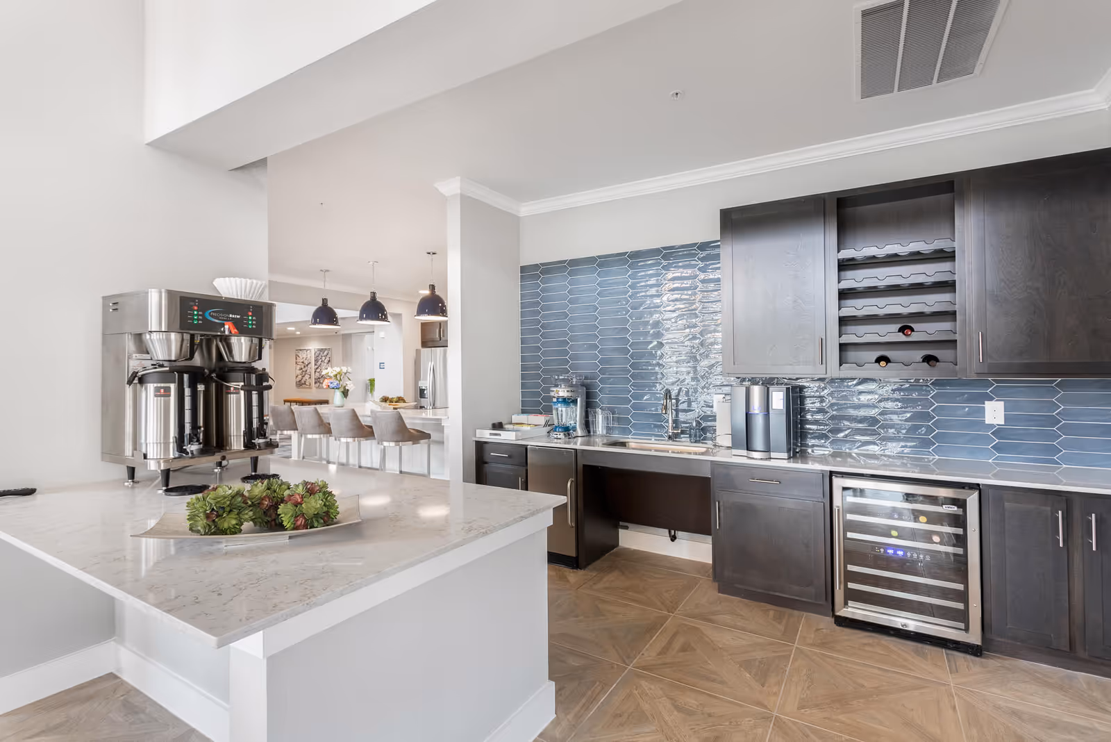 Modern kitchen area with dark wood cabinets, a blue tiled backsplash, a wine rack with several bottles, a water dispenser, coffee machine, and a stainless steel coffee brewing station on a white marble countertop. In the background, there is a dining area with a row of gray upholstered bar stools and three pendant lights hanging from the ceiling.