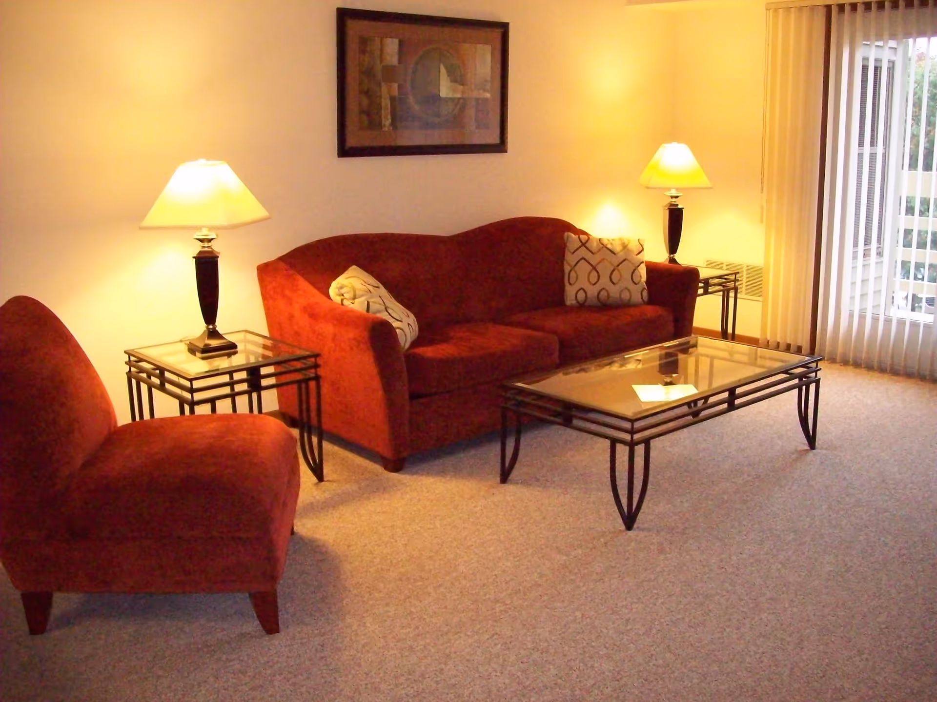 A cozy living room with a red upholstered sofa and matching chair, two glass-top side tables each with a lamp, a glass-top coffee table, a framed abstract painting on the wall, and vertical blinds covering a sliding glass door.