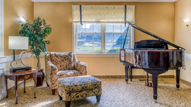 A cozy interior room with a patterned armchair and matching ottoman next to a side table with a lamp and a potted plant. A grand piano is positioned near a window with a view outside. The walls are painted a warm beige color and the floor is carpeted.