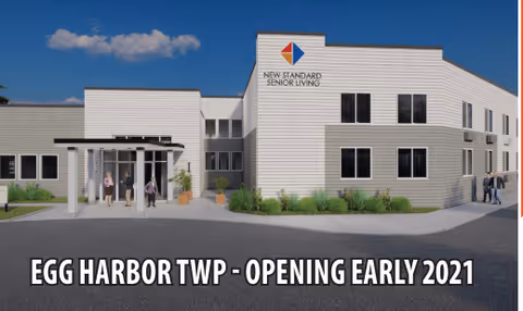 Exterior view of the New Standard Senior Living facility at Egg Harbor Township, showing a modern two-story building with white siding, multiple windows, and an entrance with a covered walkway. The sky is blue with some clouds, and there are a few people near the entrance and on the sidewalk.