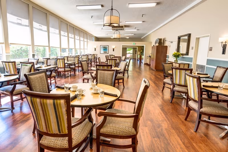 A bright and spacious dining room with multiple round wooden tables and cushioned chairs arranged neatly. The room has large windows with roller shades letting in natural light, wooden flooring, and ceiling lights. Some tables have cups, glasses, and napkins set on them. There is a doorway at the far end and a sideboard with a mirror and flowers on the right wall.