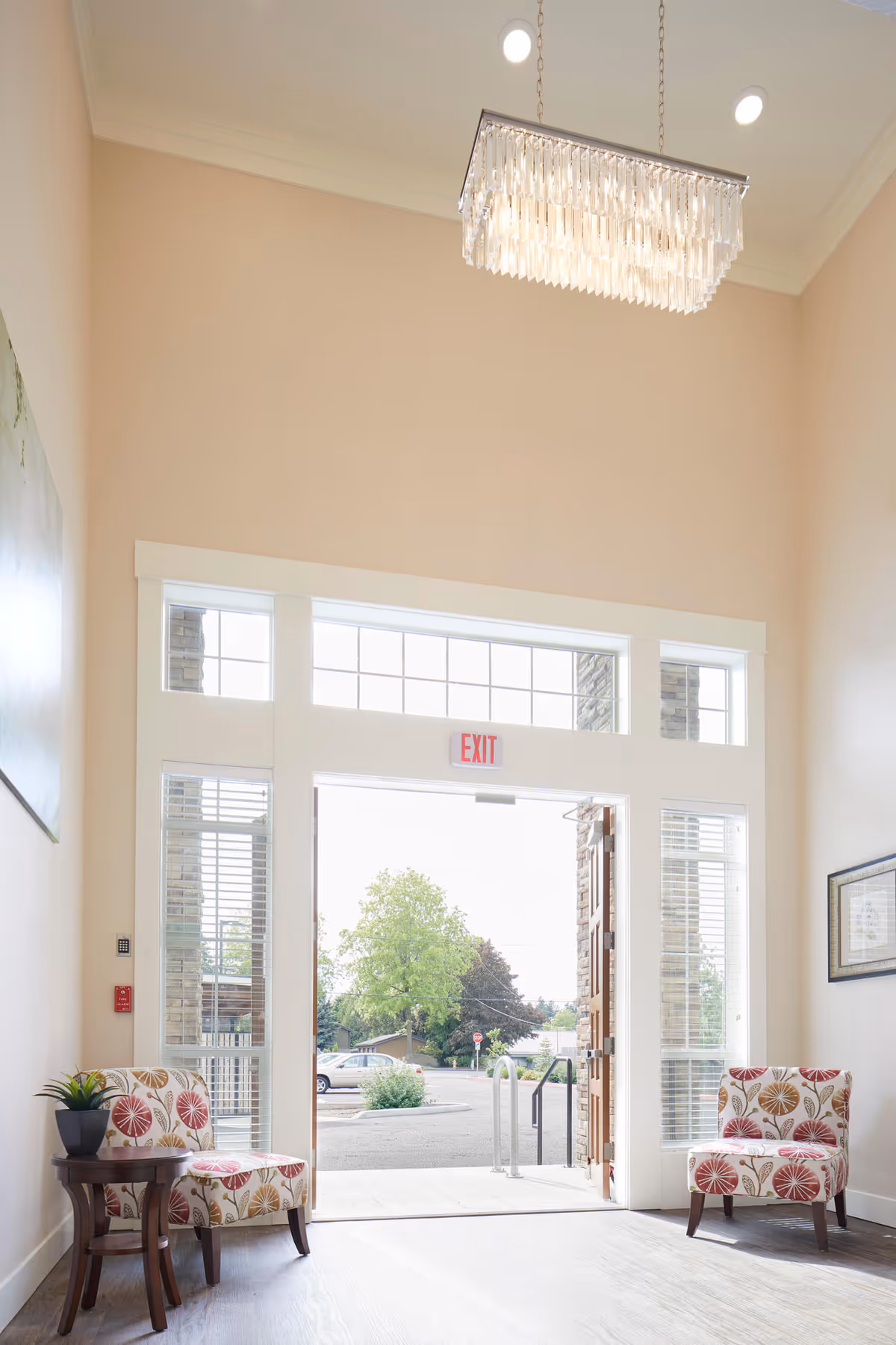 Bright entrance area of a facility with an open door leading outside. Two patterned armchairs with floral designs are placed on either side of the doorway. A small wooden side table with a potted plant is next to the left chair. Large windows surround the door, allowing natural light to fill the space. A modern rectangular chandelier hangs from the high ceiling. Outside, trees and parked cars are visible.