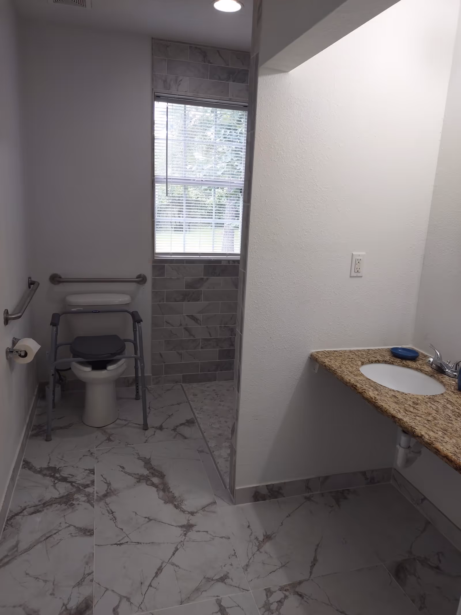 A bathroom with marble-patterned floor tiles and a walk-in shower with gray tiles. There is a toilet with a raised seat and a walker frame around it, grab bars on the walls, a toilet paper holder, and a granite countertop with a sink and faucet. A window with blinds is above the toilet.