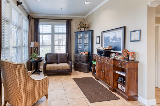 A cozy living room area with large windows covered by white blinds and brown curtains. The room features a brown cushioned loveseat, a patterned armchair, a small round side table with a lamp and plant, a dark wooden cabinet with glass doors, and a wooden sideboard with various decorative items including framed pictures and small pumpkins. The floor is tiled and there is a brown rug in front of the sideboard.