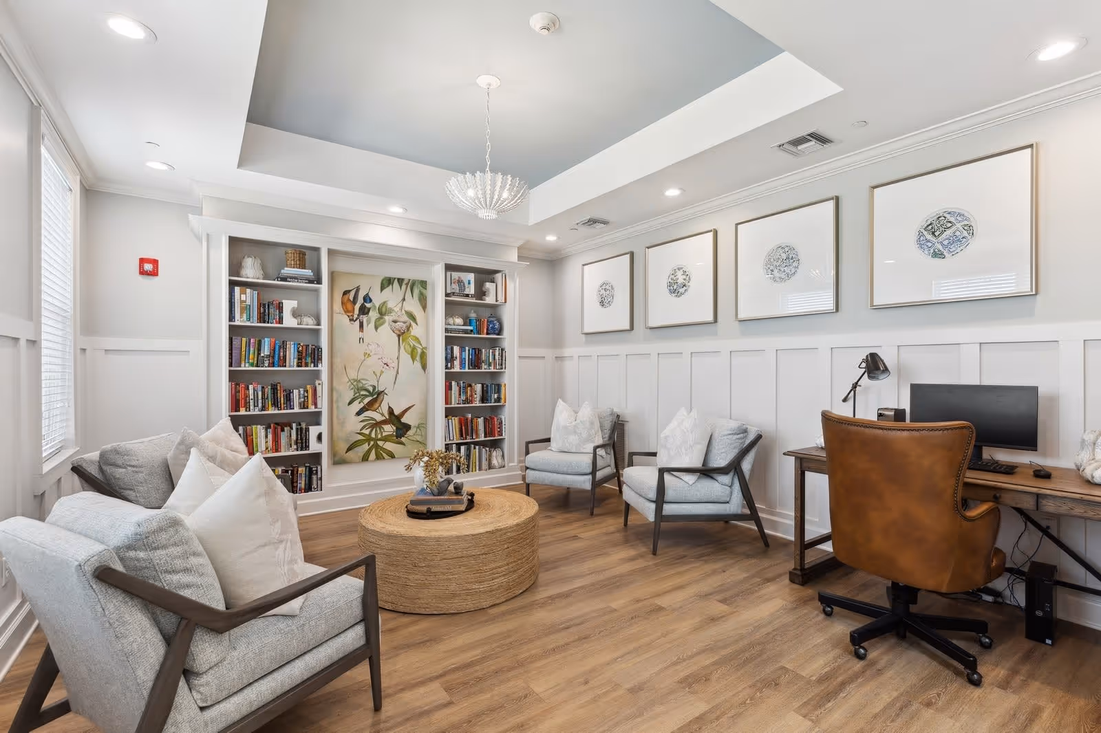 A bright and cozy living room area with light gray armchairs and white pillows arranged around a round woven coffee table. The room features built-in white bookshelves filled with books and decorative items, a large framed botanical artwork in the center, and four framed circular art pieces on the adjacent wall. There is a wooden desk with a brown leather office chair and a computer setup on the right side. The room has wood flooring, white paneled walls, and recessed ceiling lights.