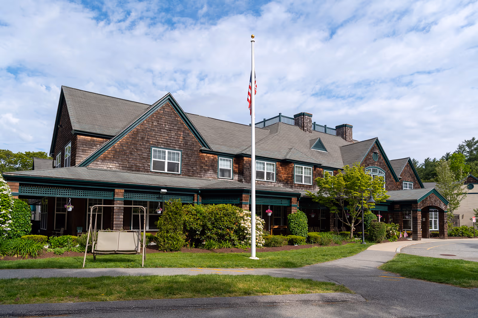Exterior view of a large two-story senior living facility building with brown shingle siding, multiple windows, and a covered porch area. There is a flagpole with an American flag in front of the building, surrounded by green bushes, trees, and a lawn. A swing bench is visible on the lawn near the sidewalk leading to the entrance.