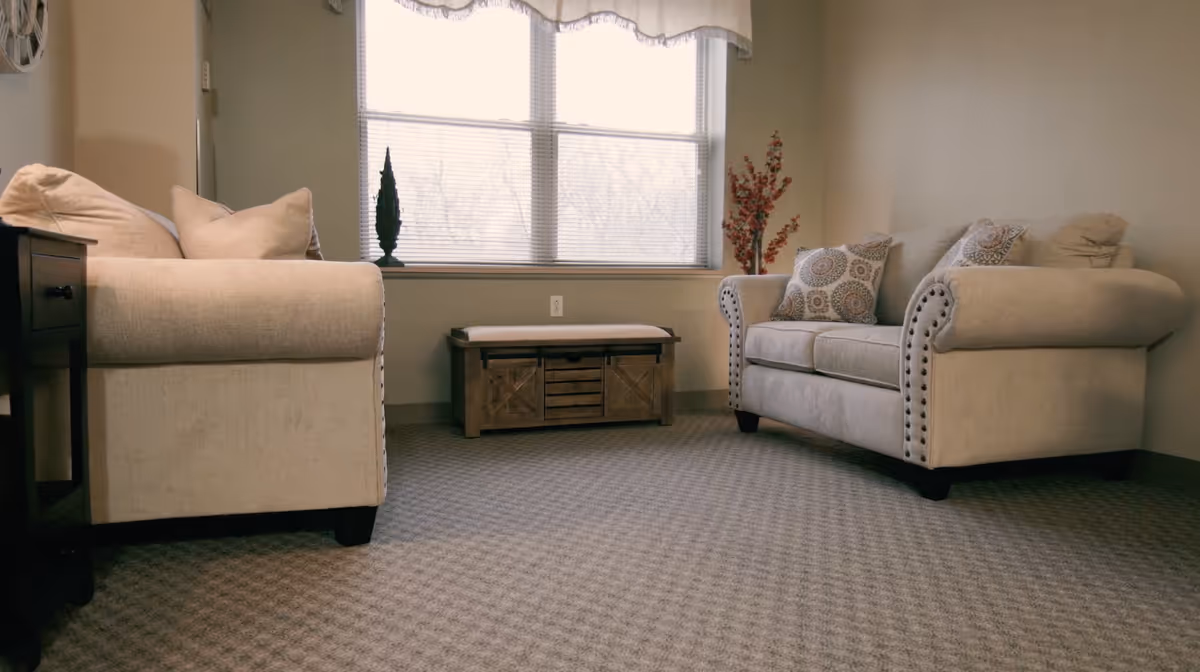 Two beige upholstered sofas facing each other in a carpeted room with a window and a small wooden bench between them.
