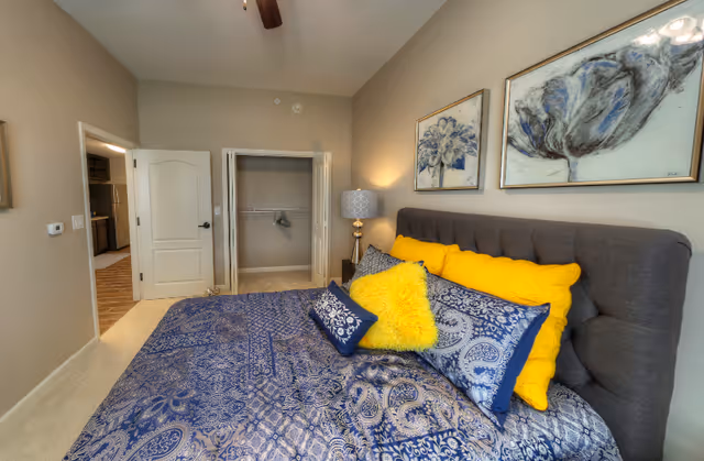 A bedroom with a large bed featuring blue and white patterned bedding and bright yellow accent pillows. The room has beige walls, two framed floral artworks above the bed, a bedside table with a lamp, an open closet with a single hanger, and a door leading to another room with a visible kitchen area.