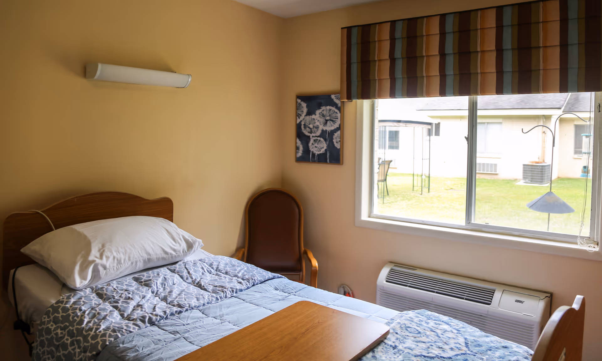 A small senior facility bedroom with a single bed, a chair, a window overlooking a courtyard, and a wall-mounted air conditioner.