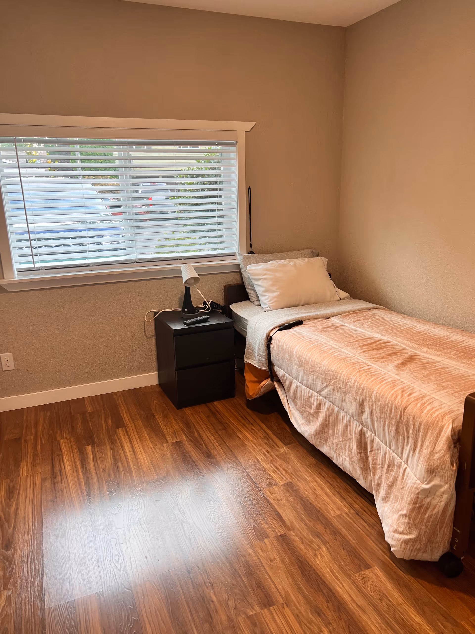 Small bedroom with a single bed, nightstand and lamp beneath a window with blinds and wood flooring.