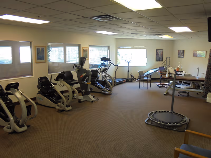 A spacious interior exercise/rehab room with recumbent bikes, treadmills, a small trampoline, and a therapy table.