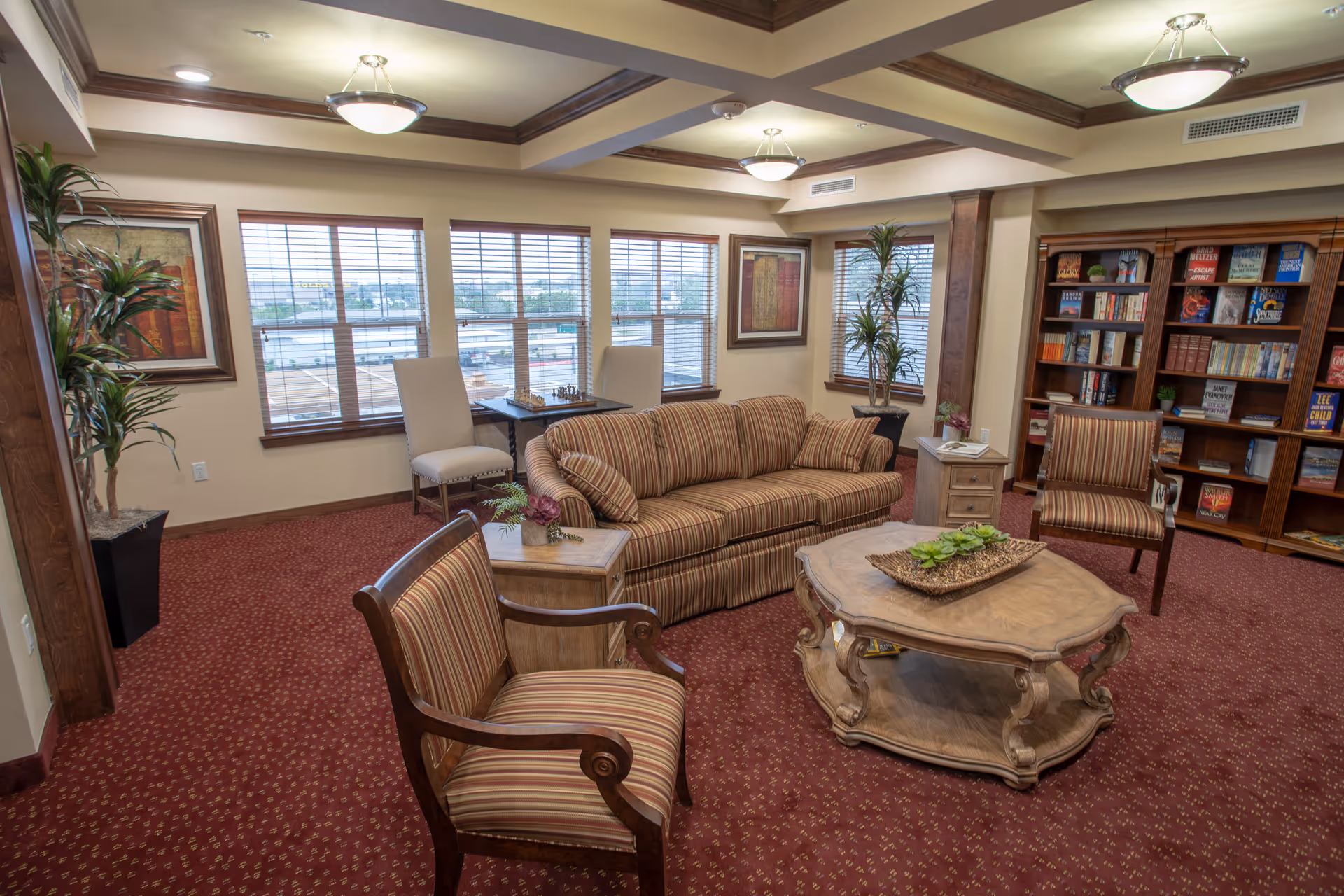 A cozy living room area with a striped sofa, two matching armchairs, a wooden coffee table with a decorative centerpiece, and a side table with a small plant. The room has large windows with blinds, framed artwork on the walls, potted plants, and a bookshelf filled with books. The carpet is red with a subtle pattern, and the ceiling features recessed lighting and wooden beams.