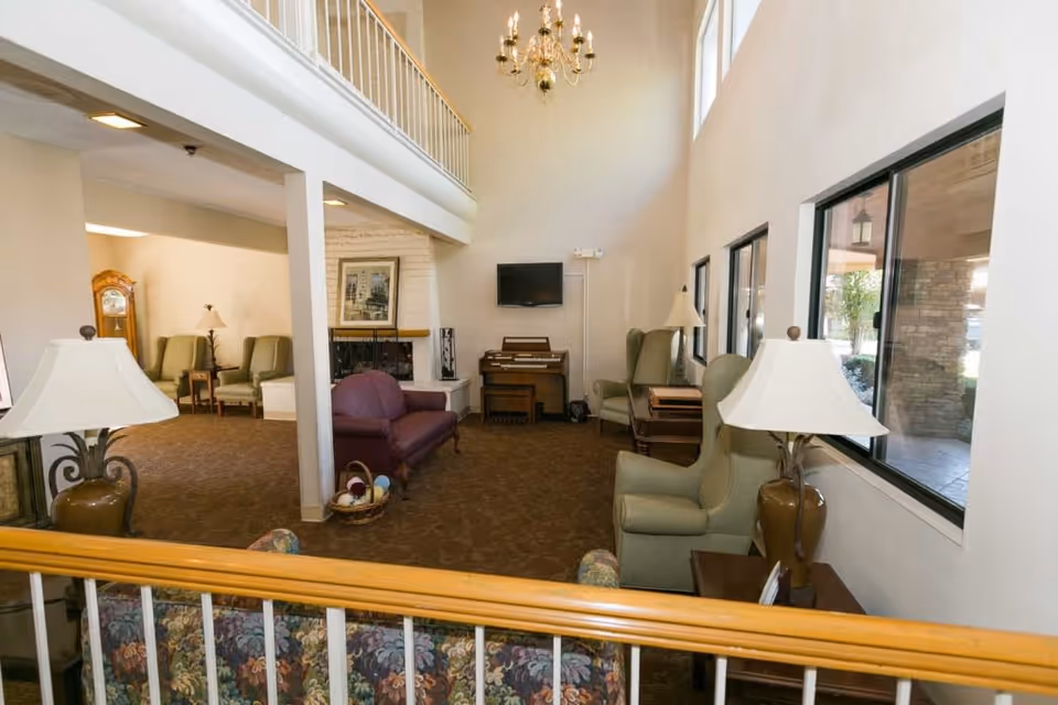 Interior view of a senior living facility common area with multiple armchairs, a purple loveseat, a fireplace, a wall-mounted TV, a piano, and large windows letting in natural light. The space has a high ceiling with a chandelier and a carpeted floor.