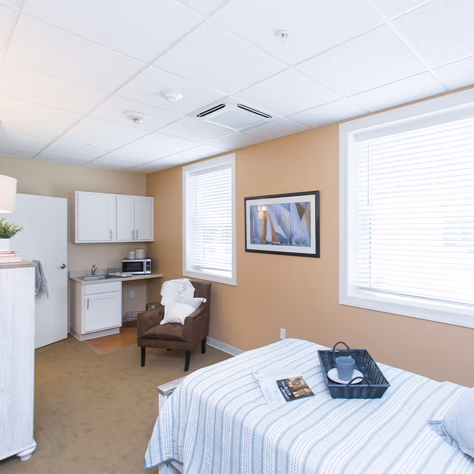 A bright and cozy senior living bedroom featuring a bed with a striped blanket, a black tray with a cup and saucer, a brown armchair with a white pillow and blanket, two large windows with white blinds, a small kitchenette with a sink, microwave, and white cabinets, and a framed abstract painting on a beige wall.