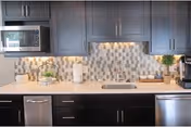 Modern kitchen interior with dark cabinets, a microwave, a stainless steel dishwasher, a sink, and a coffee maker. The backsplash features a geometric tile pattern in neutral tones.