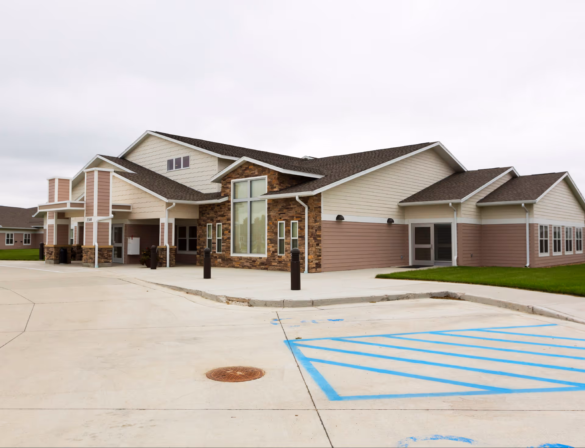 Entrance view of a single-story senior living facility with stone and siding facade, large windows, and a paved driveway with marked handicap parking.