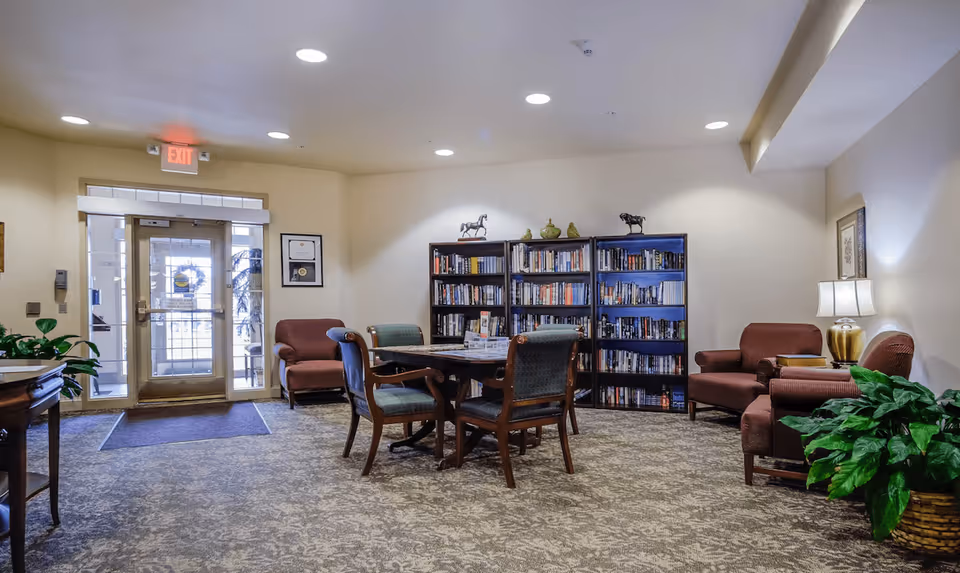 A cozy interior common area in Devonshire Retirement Village featuring a table with four chairs in the center, two bookshelves filled with books against the back wall, two armchairs on the right side with a lamp on a side table, and an exit door with glass panels on the left side. The room is carpeted and decorated with plants and framed pictures on the walls.