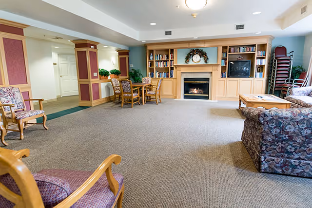 Spacious common room with upholstered chairs and sofas, a fireplace flanked by bookshelves, and a table with chairs.