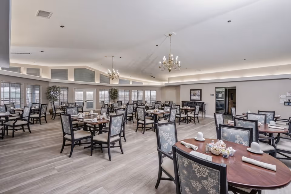 Spacious dining room with multiple wooden tables and cushioned chairs arranged neatly. Each table is set with napkins, cups, and condiments. Large windows line the walls, allowing natural light to fill the room. The ceiling features elegant chandeliers and recessed lighting. The floor is covered with light-colored wood laminate, and there are a couple of potted plants near the windows.