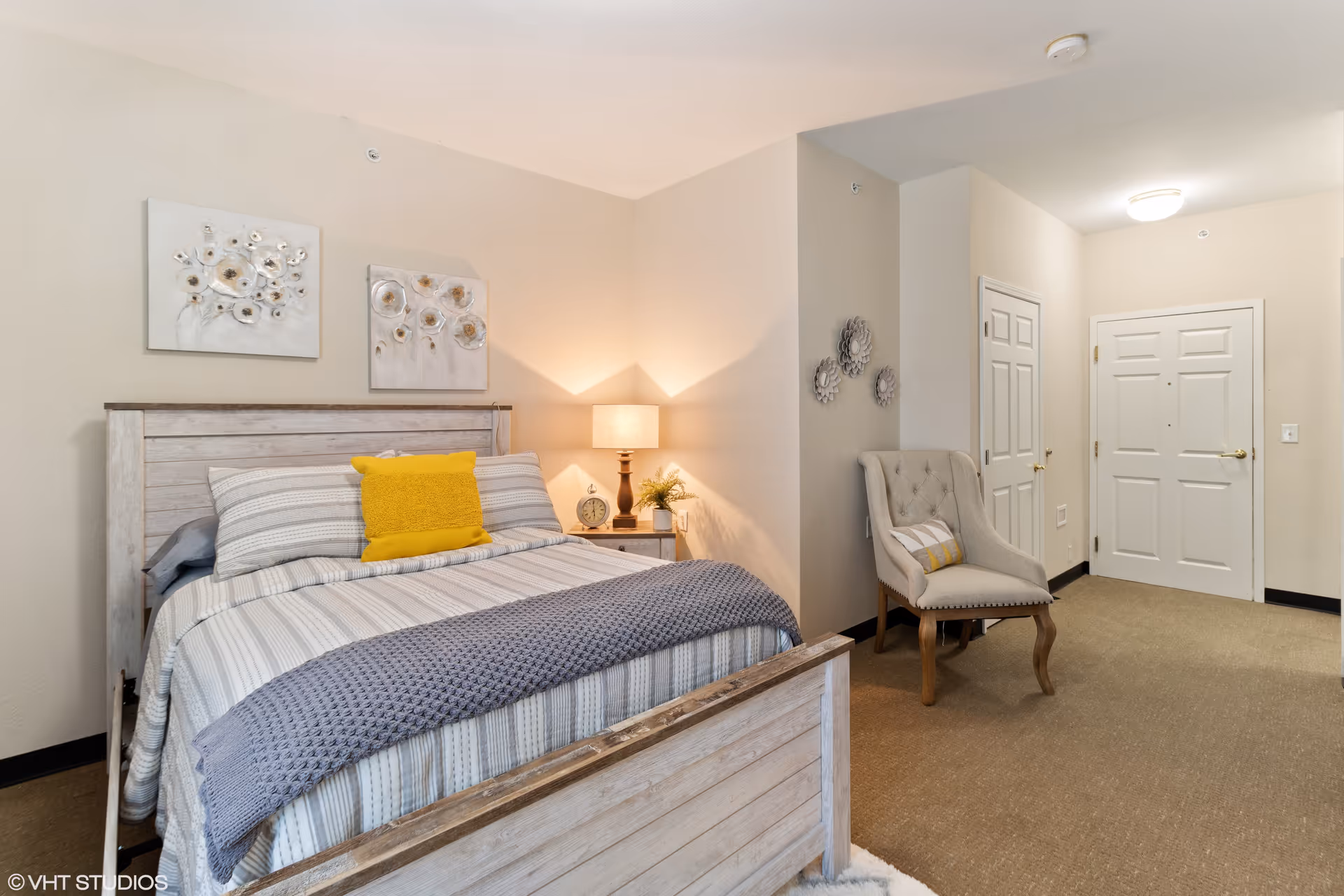 A cozy bedroom featuring a wooden bed with striped bedding, a yellow accent pillow, and a gray knitted throw blanket. A wooden nightstand with a lamp, clock, and small plant is beside the bed. On the wall above the bed are two floral paintings. A beige upholstered chair with a decorative pillow sits near the entrance door, with three decorative wall hangings above it. The room has beige walls and carpeted floor.