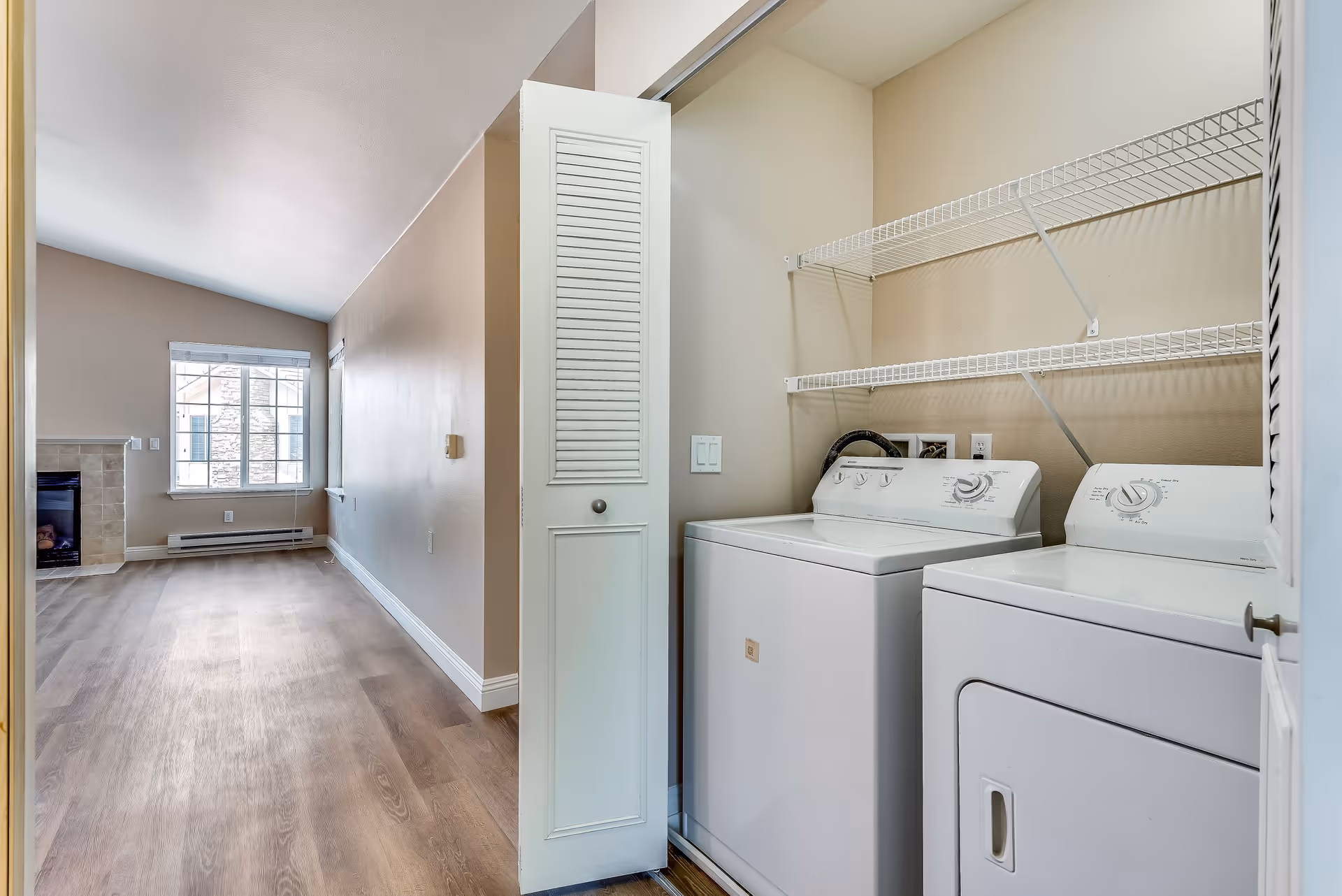Interior view of a senior living facility showing a laundry area with a washing machine and dryer behind white louvered doors. The room has beige walls and wooden flooring. In the background, there is a living area with a window and a fireplace.