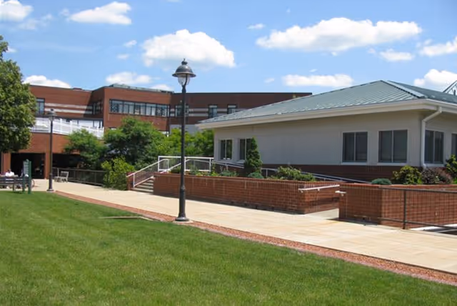 Outdoor view of a senior living facility with a paved walkway, green lawn, a lamppost, and a building with a green roof and brick walls under a blue sky with scattered clouds.