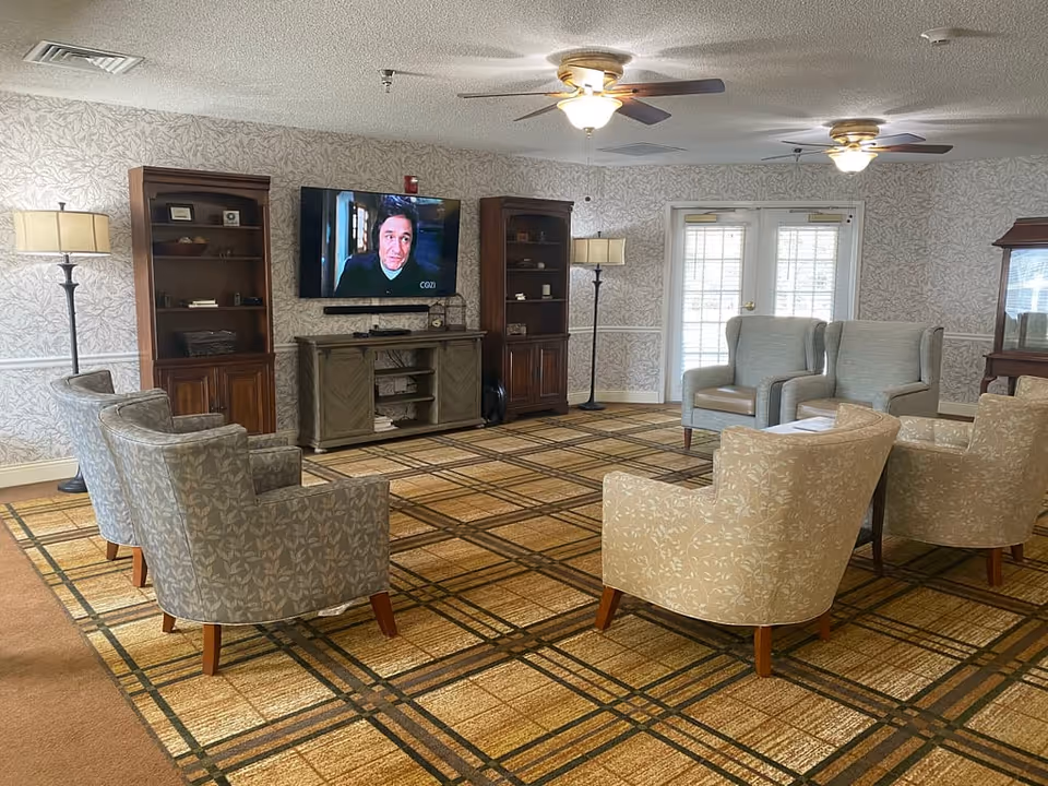 A bright common living room with upholstered armchairs arranged around a wall-mounted television, bookshelves, floor lamps, and ceiling fans.