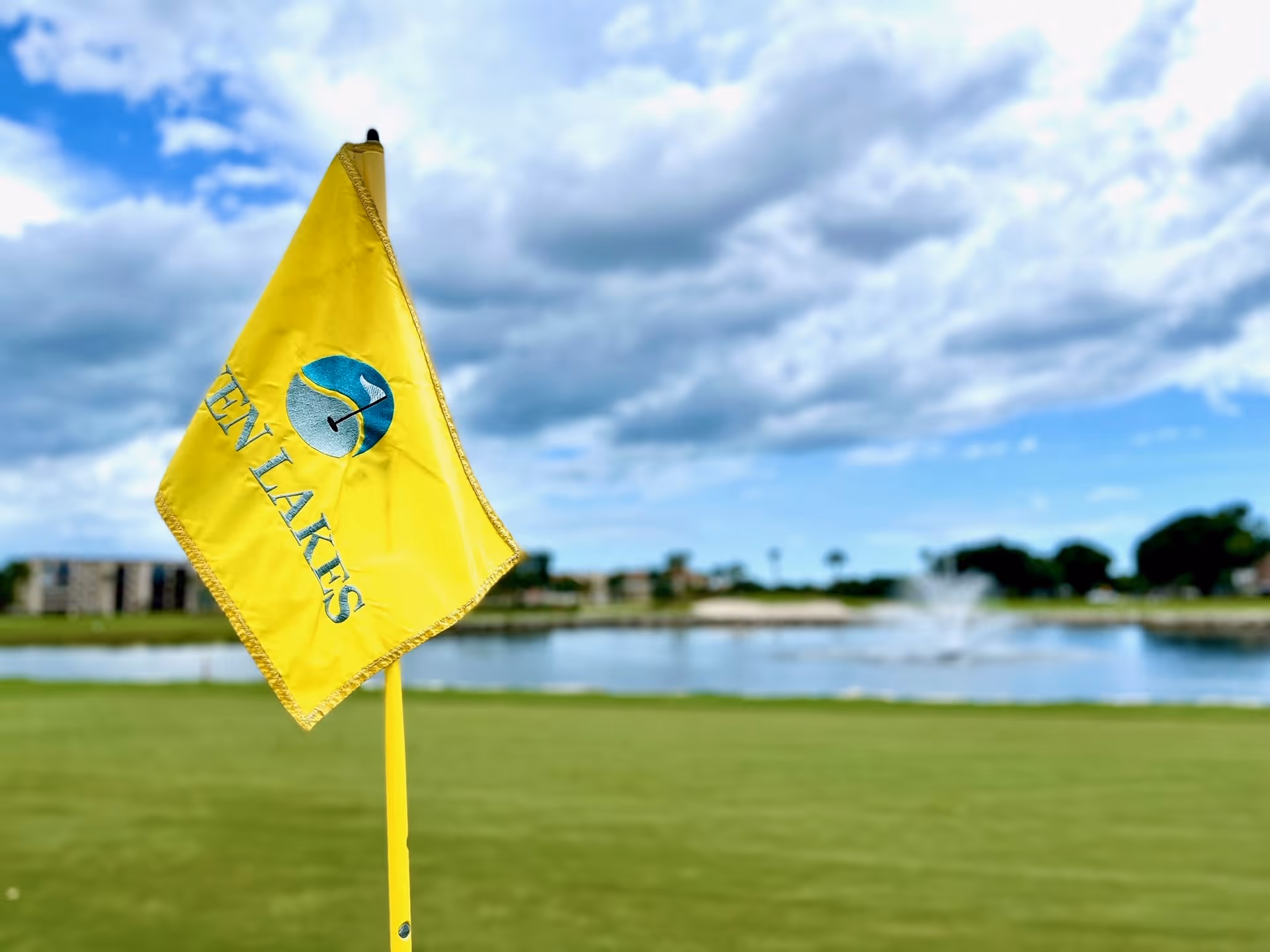 Yellow golf flag with the text 'Seven Lakes' on a golf course green, with a water fountain and buildings in the background under a partly cloudy sky.