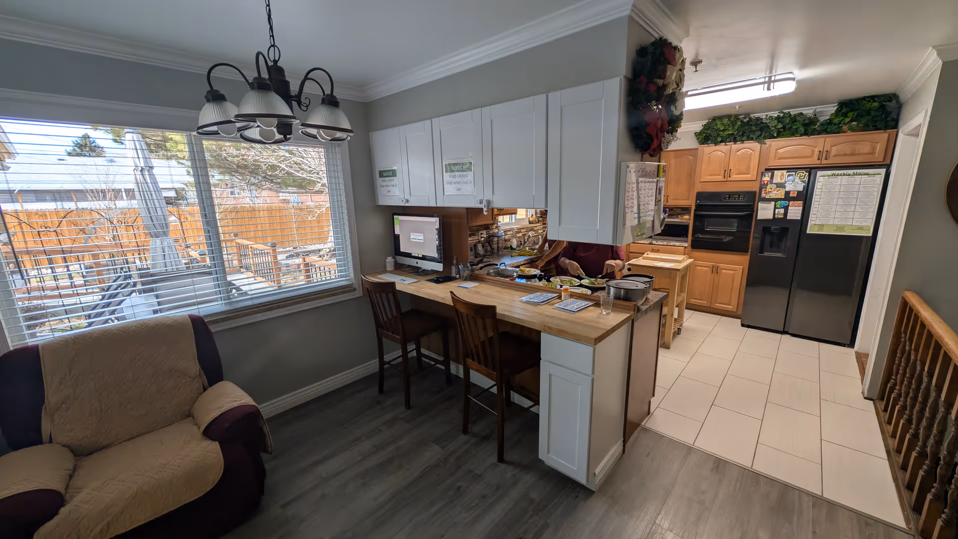 Interior view of a kitchen and dining area in an assisted living facility. The kitchen has wooden cabinets, a black refrigerator with magnets and notes, a double oven, and a countertop with two wooden chairs. There is a computer on the counter and some papers and dishes. A large window with blinds shows an outdoor patio area with tables and umbrellas. A cushioned armchair is positioned near the window, and a hanging light fixture is above the dining area.