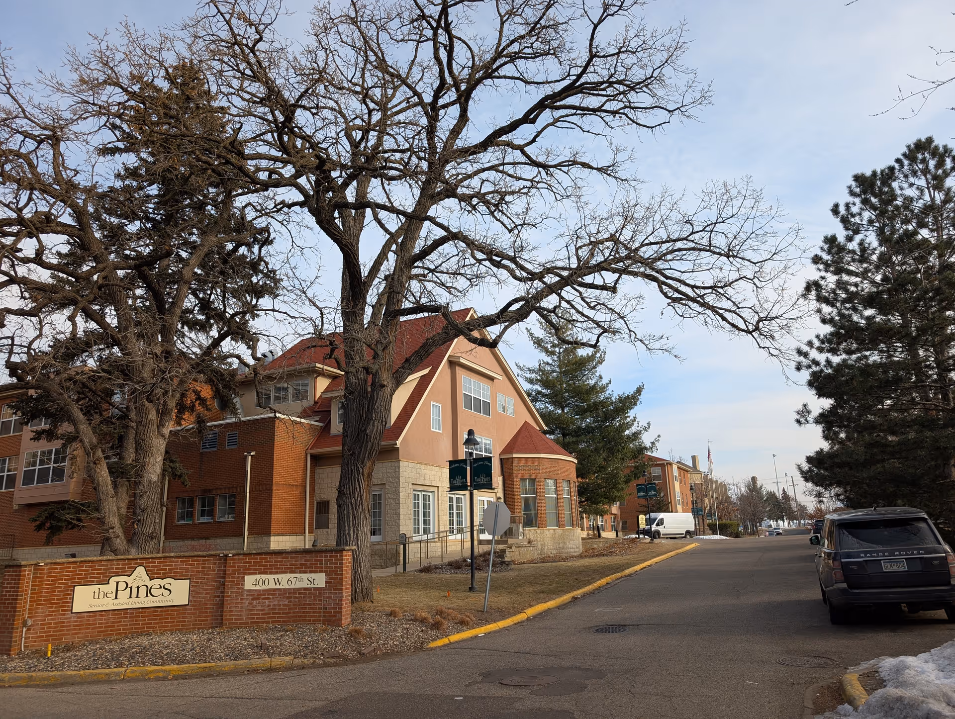 Front exterior of The Pines senior living building with a brick sign, leafless trees, and a street with parked cars.