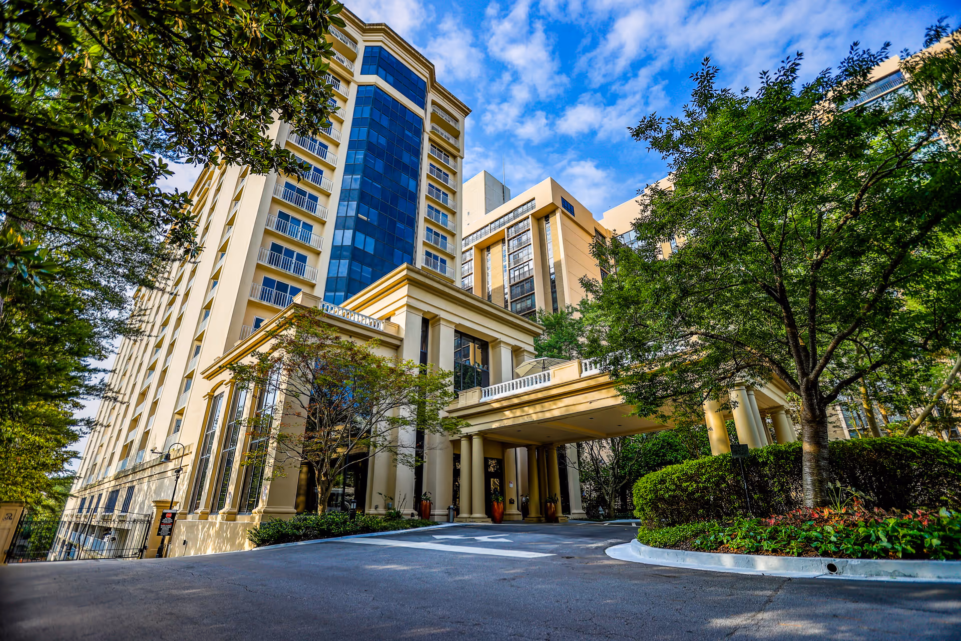 Exterior view of Renaissance on Peachtree, a multi-story building with large windows and a covered entrance. The building is surrounded by trees and greenery under a partly cloudy blue sky.
