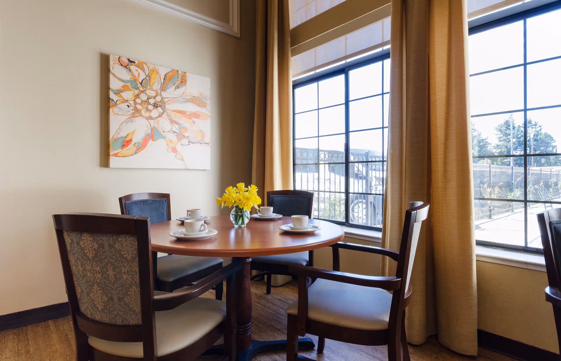 A small round wooden dining table set with four chairs, each with cushioned seats and patterned backs. The table has four place settings with white cups and saucers, and a small vase with yellow flowers in the center. The room has large windows with beige curtains allowing natural light to enter, and a colorful abstract flower painting hangs on the wall.