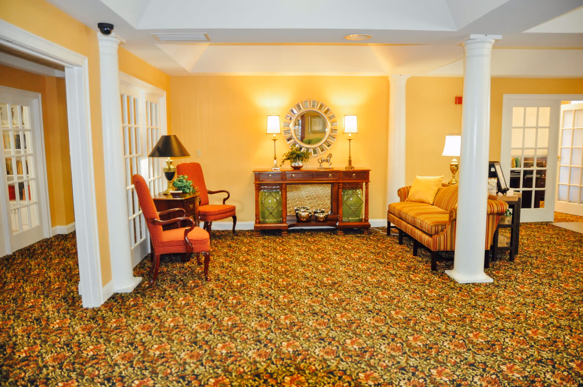 A cozy sitting area with patterned carpet, two red upholstered armchairs on the left, a wooden side table with a black lamp and plant, a wooden console table with decorative items and a round mirror above it on the back wall, and a striped sofa with a yellow pillow on the right. The walls are painted yellow and there are white columns and French doors visible.