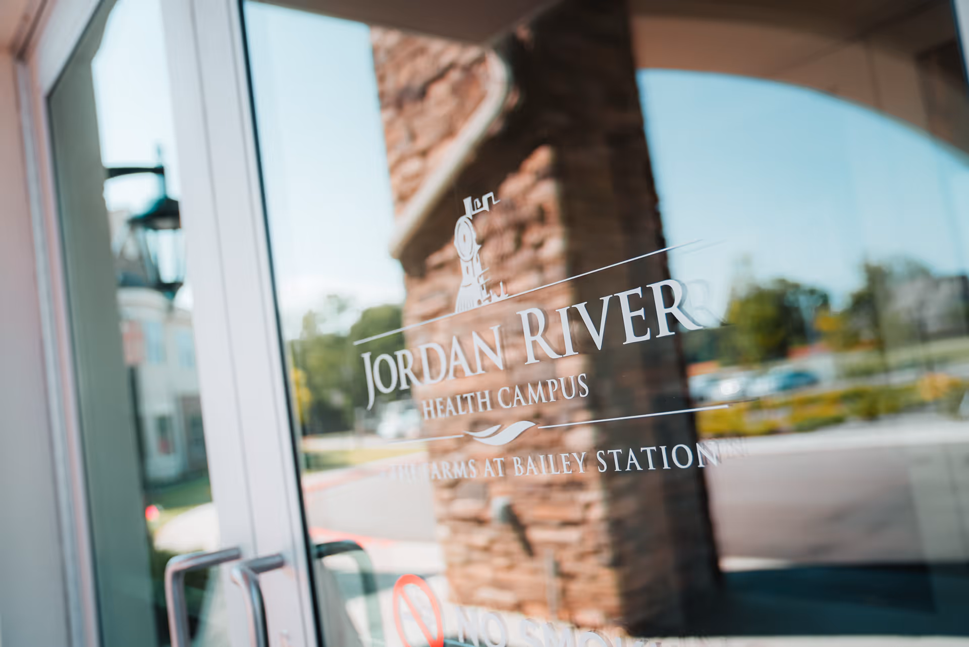 Glass entrance door with the logo and text 'Jordan River Health Campus The Arms at Bailey Station' printed on it, reflecting a stone pillar and outdoor surroundings including trees and parked cars.