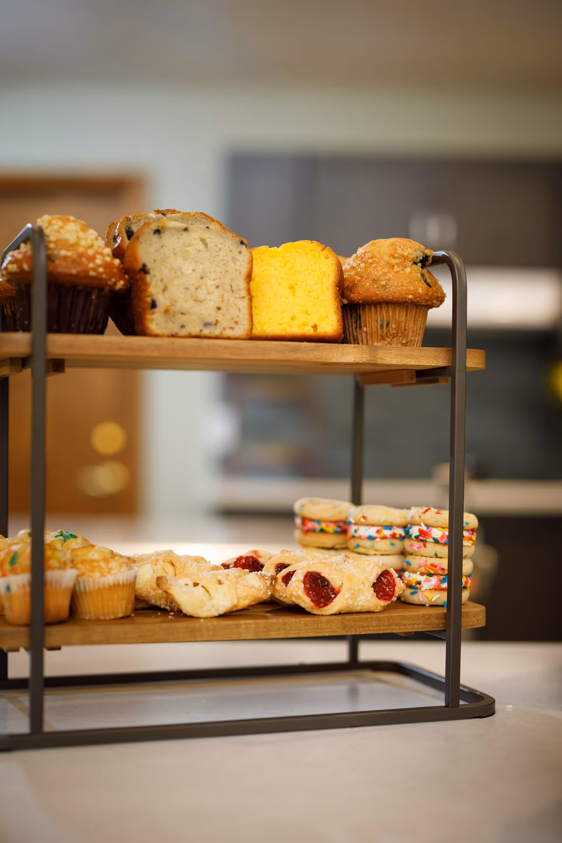 Two-tiered wooden and metal stand displaying assorted baked goods including muffins, slices of bread, pastries with fruit filling, and sandwich cookies with colorful sprinkles in a kitchen setting.