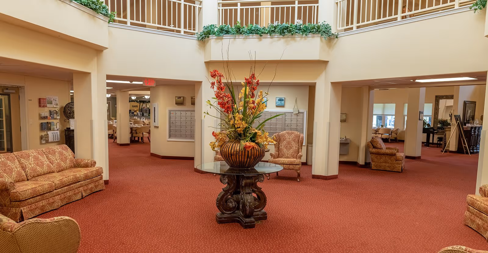 Interior view of a senior living facility lobby with red carpet, beige walls, and multiple seating areas with patterned sofas and chairs. A round glass table with a large floral arrangement is centered in the space. Mailboxes are visible on the far wall, and the area is well-lit with natural and artificial light.