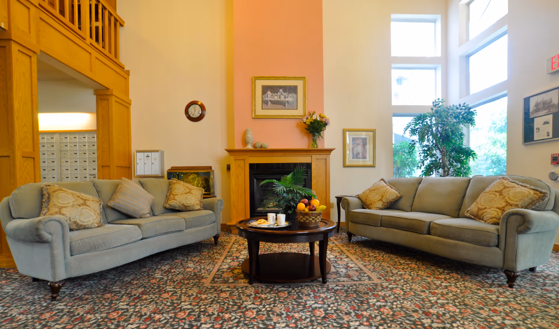 A cozy senior living facility common area with two light gray sofas facing each other, adorned with patterned cushions. A round wooden coffee table sits in the center with a basket of fruit and two cups. Behind the table is a wooden fireplace with a pink accent wall, decorated with framed pictures and a vase of flowers. Large windows allow natural light to brighten the room, and a green plant is placed near the window. The floor is covered with a patterned carpet, and wooden architectural details are visible on the left side.